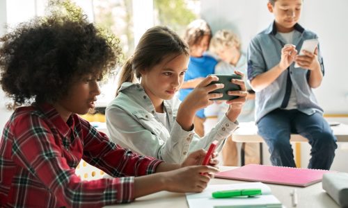 Diverse multiethnic kids school students using smartphones in classroom. Diverse multiethnic kids students using smartphones in classroom. Multicultural children holding devices having fun with mobile phones apps playing games and checking social media at school.